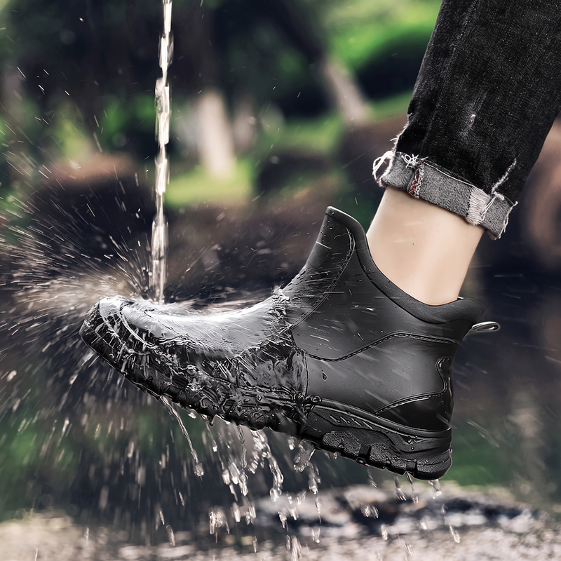 A black ankle rain boot being splashed with water while worn on a person's foot, with cropped dark jeans visible above the ankle.