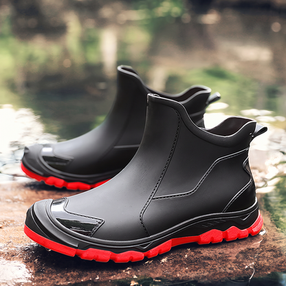 A pair of black ankle rain boots with red soles sitting on a wet rock near water.