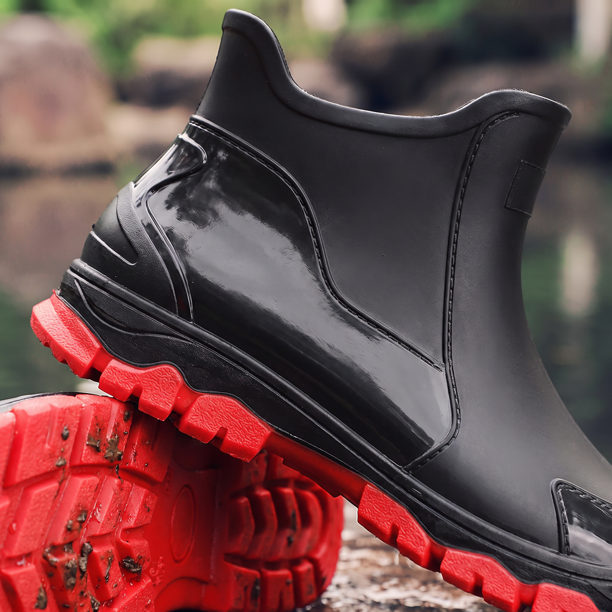 A pair of black ankle rain boots with red soles, one worn on a foot and one held up by a hand, near a wet rocky area with water in the background, close-up view