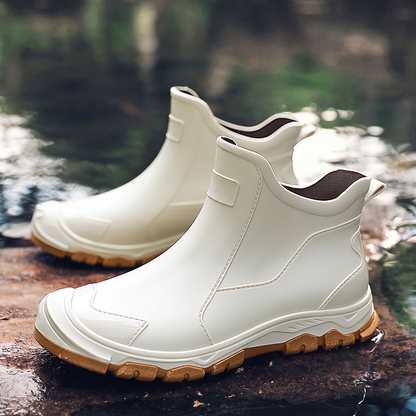A pair of cream colored ankle rain boots with tan soles sitting on a wet rock near water.