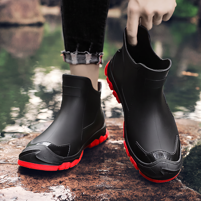 A pair of black ankle rain boots with red soles, one worn on a foot and one held up by a hand, near a wet rocky area with water in the background.