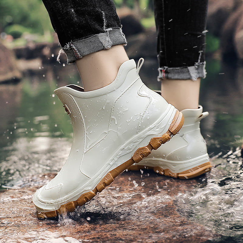 A person wearing cuffed black jeans steps through a wet rocky stream wearing white ankle rain boots with a brown ridged sole.