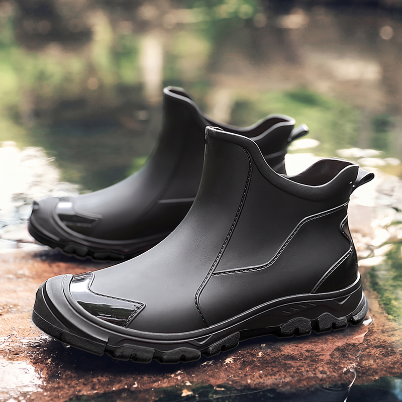 A pair of black ankle rain boots with a side logo patch displayed on a wet stone surface by water.