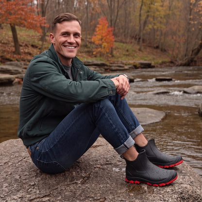 Mann sitzt am Fluss, trägt schwarze Regenstiefel mit roter Sohle | Chaps Herrenmode