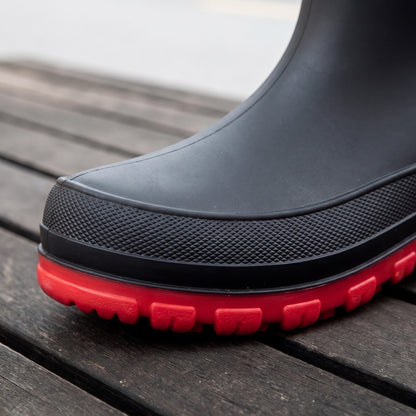 Schwarzer Gummistiefel mit roter Profilsohle, Nahaufnahme der Zehenpartie auf Holzboden | Chaps Herrenmode
